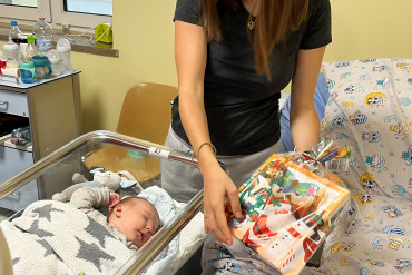 Na zdjęciu śpiące niemowlę, nad nim stoi mama trzymająca w rękach prezent
