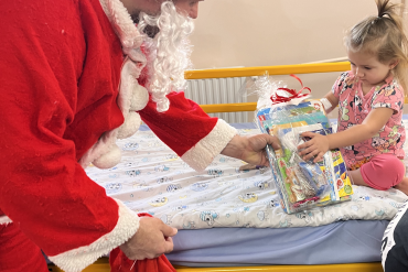 Mała dziewczynka siedzi na łóżku, na przeciwko niej pochyla się św. Mikołaj wręczając jej prezent.