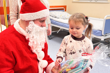 Św. Mikołaj kuca przy małej dziewczynce wręczając mu prezent, za nimi stoi aniołek.