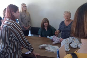 Grupa kobiet zebrana wokół stołu, na stole leży lalka - niemowlę.